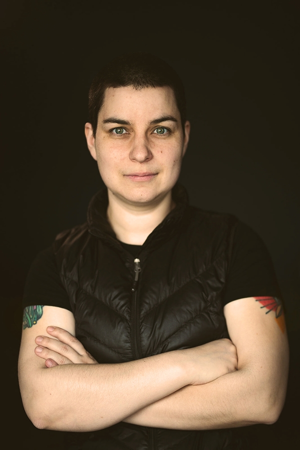 Max stands with arms crossed in front of a dark background. They have short cropped black hair, light skin and green eyes. They are wearing a black vest over a black short sleeved shirt and have tattoos visible on their biceps. 